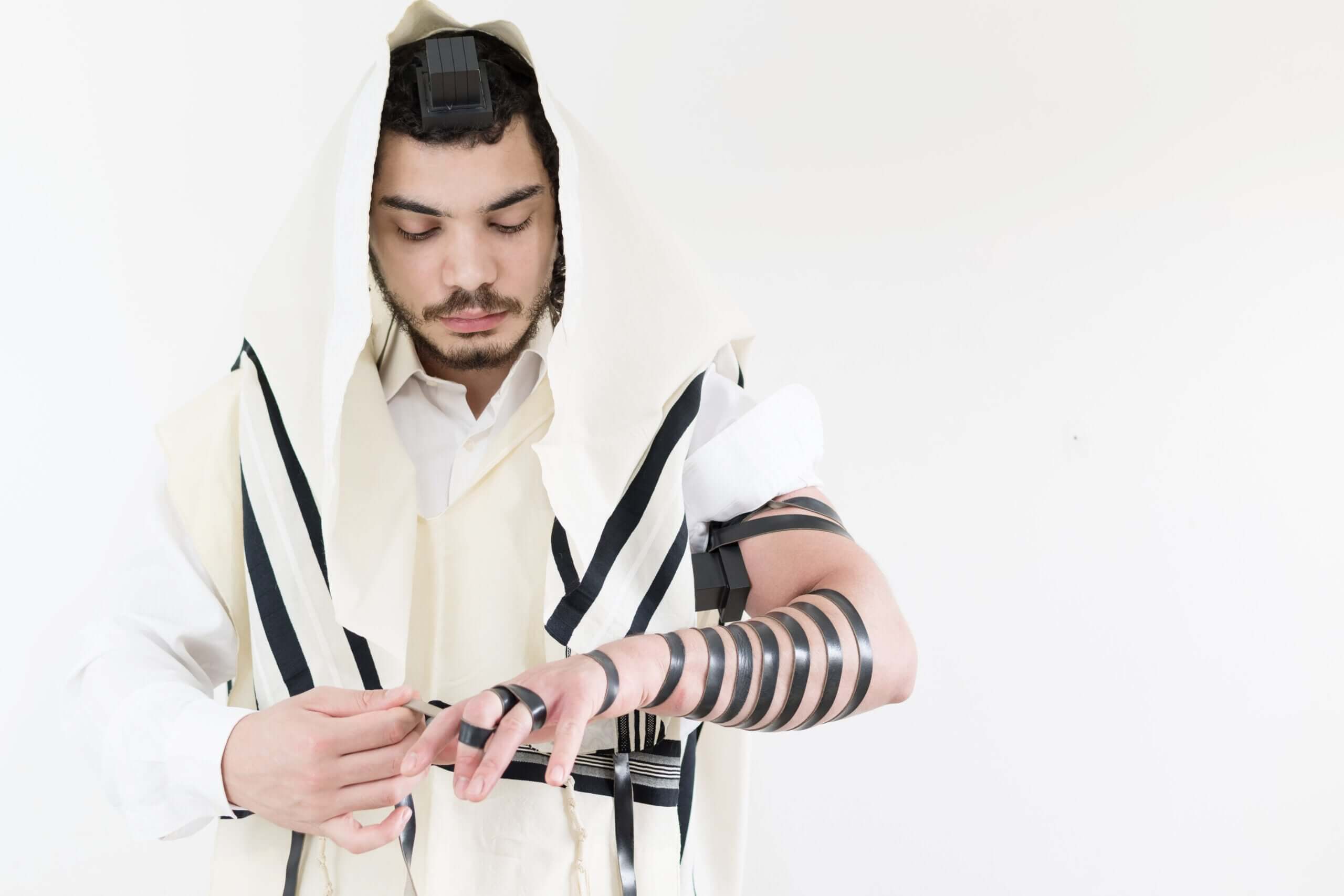 Jewish man wrapping tefillin around his fingers. Has the tefillin on his head and wearing a tallit.