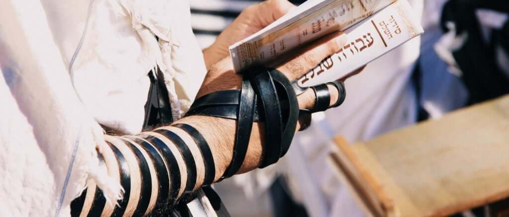 Man praying from a siddur wrapped in tefillin, focused on how it is wrapped around his hand.